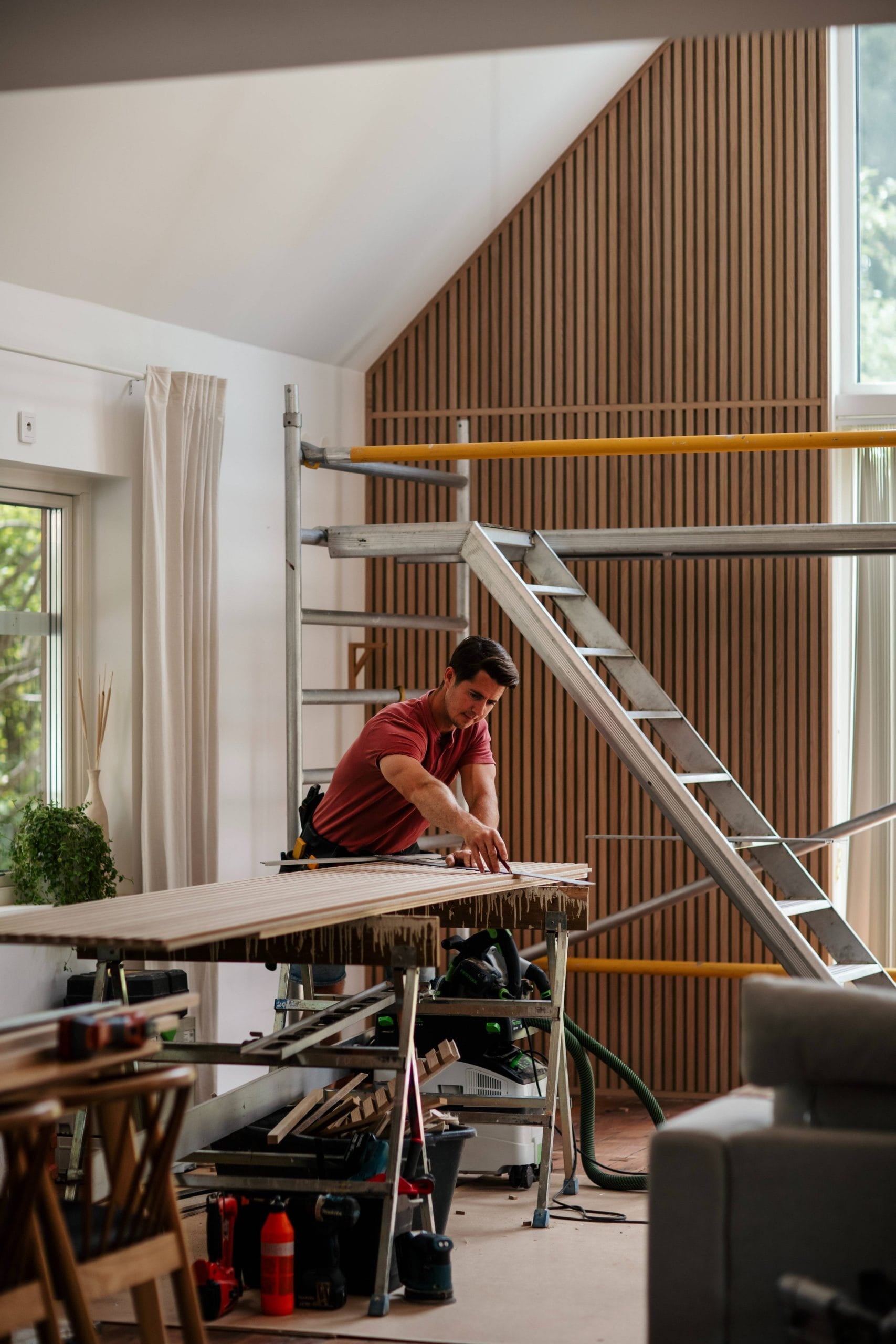 Un homme mesure une planche de bois sur un établi dans une pièce moderne, partiellement rénovée, avec des échafaudages et de grandes fenêtres, suggérant linstallation de panneaux acoustiques.